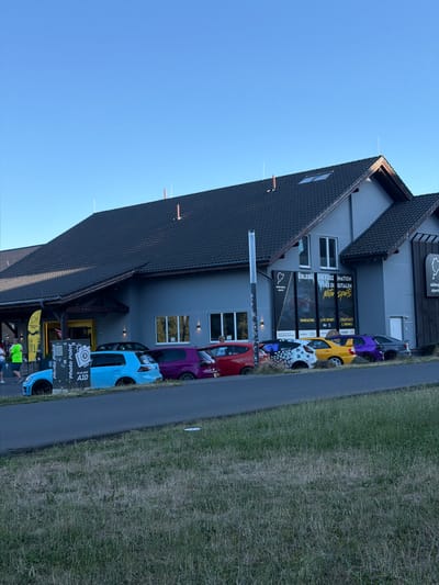 The image depicts the exterior of a building in Nürburg, Germany, with several cars parked in front of it. The building is a two-story structure with a dark-tiled roof and light-colored walls. There are several windows and entrances visible. A prominent sign with the words "ERLEBE DIE FASZINATION DES DIGITALEN Motor Sports" is displayed on the building. In front of the building, a row of cars are parked, including a bright blue Volkswagen Golf, a purple Volkswagen Golf, a red Volkswagen Golf, a yellow Volkswagen Golf, a white Volkswagen Golf with a distinctive graphic design, and a dark-colored sedan. A sign for "Photo Point at car park A10" is also visible near the blue car. The area in front of the building appears to be a parking lot, with a road and a grassy embankment in the foreground. The sky is clear and blue, suggesting it is daytime. A few people are visible in the background, near the entrance of the building. The overall scene suggests a motorsports-related venue or event.