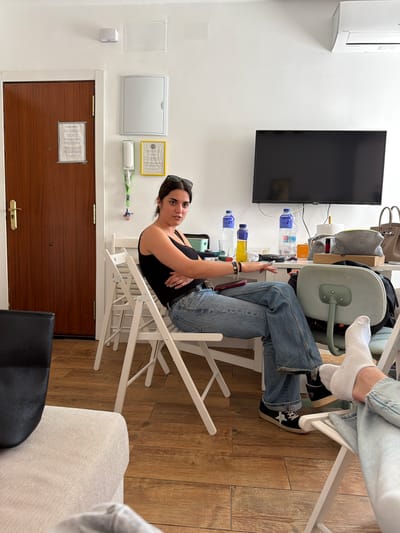 The image, captured from a slightly elevated perspective and rotated 90 degrees clockwise, depicts an indoor room with two individuals. In the foreground, a young woman with dark hair, wearing a black tank top and blue jeans, sits on a white folding chair with her legs crossed. She has sunglasses perched on her head and multiple bracelets on her left wrist. She is looking directly at the camera with a neutral expression. Her feet are clad in black sneakers with white laces and a white star logo. Another person's lower legs and feet, wearing light blue jeans and white socks, are visible in the top right corner, with one foot propped on a gray office chair.

The room features white walls and a light brown wooden floor. A wooden desk or table is positioned along one wall, laden with items including several plastic water bottles (some containing blue-capped water, one with a yellow liquid), a gray handbag, a black bag, a roll of paper towels, and a phone with a red case. Above the desk, a black flat-screen television is mounted on the wall, and to its left, a white air conditioning unit is also mounted high on the wall.

To the left of the woman, a dark brown wooden door with a gold handle is visible, featuring a white paper notice taped to its surface (text illegible). Above this door, various wall fixtures include a white rectangular panel, a grey rectangular box, a yellow-framed document (text illegible), and a white device with a green strap attached. Portions of a light-colored sofa/armchair and a black chair are seen in the lower right. The scene conveys a casual, relaxed atmosphere, likely taking place during the daytime.