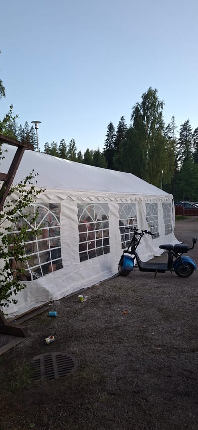 In Salpakangas, Finland, a blue electric scooter is parked on a gravel surface next to a white tent. The tent has clear windows, and people are visible inside. Scattered on the ground around the tent are empty cups and cans, indicating a gathering or event has taken place. The background features trees and a clear sky, suggesting an outdoor setting.