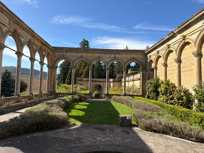 The image captures a formal garden within an architectural framework of stone arches and colonnades. The location is a courtyard in the city of Malvern Hills, United Kingdom. The garden features manicured hedges, a rectangular lawn, and stone pathways. The stonework appears weathered with age. In the background are additional stone structures and verdant trees. The sky is blue with wispy clouds. There are no people visible in the image.