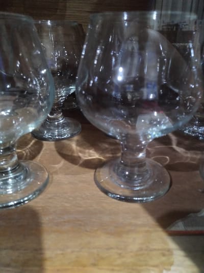 The image displays several clear glass goblets or stemmed glasses lying on their sides on a wooden surface. The glassware, appearing empty, is arranged diagonally across the frame. Their transparent nature causes strong reflections and refractions of light, creating bright spots and intricate patterns both on the glass surfaces and on the surrounding wooden table.

The wooden surface exhibits a light to medium brown tone with a visible grain, contributing to the reflections and shadows cast by the glasses. The background is out of focus, further emphasizing the glassware and the play of light. No specific activity is occurring; the glasses are static, suggesting they are either stored or recently used.

There are no people or visible text in the image. The scene is set indoors, likely on a table or shelf. The strong highlights indicate either bright indoor lighting or sunlight entering through a window. The overall impression is a detailed study of light, glass, and wood, captured in Gampongbaro, Indonesia.