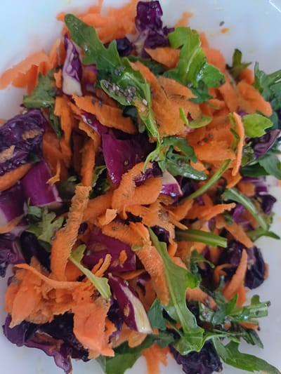 A close-up, overhead shot shows a colorful salad in a white bowl. The salad is composed of shredded carrots, dark purple cabbage, and arugula greens. Tiny black seeds, possibly chia seeds, are scattered throughout the salad. The lighting is bright and natural, suggesting the photo was taken indoors during the day. There is no indication of the location being in the city of Чёрновский, Russia.