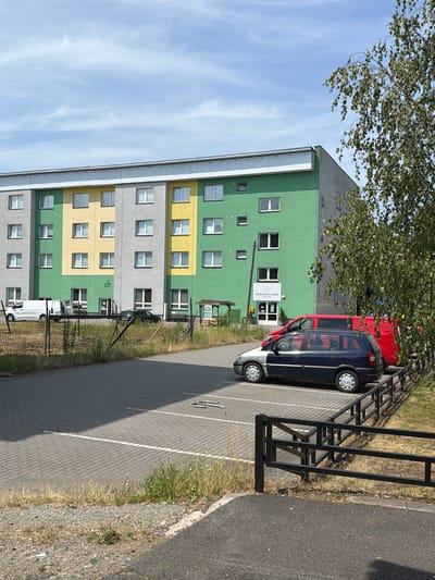 This image shows a three-story apartment building with a multi-colored facade in gray, yellow, and green. The building has numerous windows on each floor. In front of the building is a parking lot paved with interlocking bricks, with white parking lines. Several vehicles are parked in the lot, including a dark blue minivan, a red van, and some white vans closer to the building. A black metal fence separates the parking lot from a grassy area. There is a tree with green leaves in the foreground, partially obscuring the right side of the building. A sign on the building reads "Alzheimer Home" with a logo. The sky is blue with scattered clouds. The image appears to be taken on a sunny day. The specific location is the Alzheimer Home in Český Těšín, Czechia.