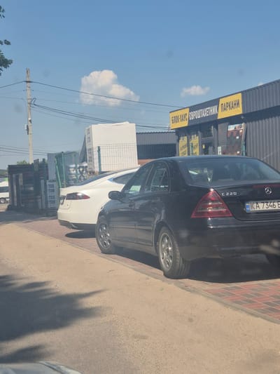 The image shows a street scene with two cars, a white Tesla and a black Mercedes-Benz, parked on a brick-patterned sidewalk next to a dirt road. In the background, there is a building with a sign in Ukrainian that reads "БЛОК-ХАУС ЄВРОШТАКЕТНИК ПАРКАНИ". There are also stacks of white building materials, possibly insulation panels, on pallets behind a green fence. The sky is blue with a few scattered clouds, and the sun is casting shadows on the ground. The overall impression is a commercial area, possibly a building materials supplier, on a sunny day in Pidhaitsi, Ukraine.