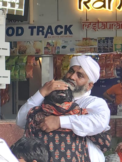 An elderly man wearing a white turban and a white shirt is embracing another person who is mostly obscured by the man's arms and a patterned shawl. The man has a grey beard and is looking down. A ring with a small stone is visible on his left hand. They are standing in front of a stall that displays various food and drink items, including packaged snacks and milk cartons. Above the stall, a sign reads "FOOD TRACK" in large blue letters, with smaller text below indicating "AN IRCTC INITIATIVE". To the right, a neon sign glows with the word "Ran" in yellow, and a smaller red sign below it reads "restaurant". The scene appears to be outdoors, likely in a public area like a train station, given the "IRCTC" branding, which is associated with Indian Railways. The overall impression is one of an emotional embrace in a bustling commercial environment.