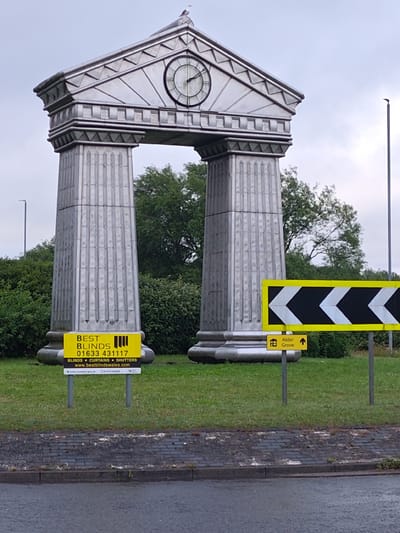 The image shows a monument with classical architectural elements, specifically two large fluted columns supporting an entablature with a clock face and a bird on top. The monument is situated in a grassy area, with trees visible in the background. In the foreground, there are several signs. One is a yellow rectangular sign for "BEST BLINDS" with a phone number and website address. Another sign is a directional sign with yellow and black chevrons indicating a turn, and below it, a smaller yellow sign with a house icon and the text "Alder Grove" pointing upwards. The overall setting appears to be a roundabout or junction in a suburban area. Based on the "01633" area code on the "Best Blinds" sign, this location is likely in or near Newport, United Kingdom, as 01633 is the telephone dialling code for Newport and surrounding areas.