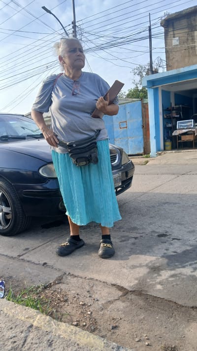An elderly woman stands beside a dark blue car in a residential area in Barcelona, Venezuela. She has white hair, is wearing a light grey t-shirt, a pleated light blue skirt, black socks and Croc-style shoes with gold embellishments. A pair of glasses hangs from her shirt collar. She is holding a clipboard in her left hand and rests her right hand on the car. A black bag is slung across her body. Behind the car is a blue gate and the corner of a building with blue trim. Overhead, a tangle of power lines is visible. The road is concrete and shows signs of wear. The license plate of the car is partially visible, reading "16GED".