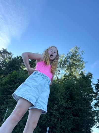 A young female with blonde hair, wearing a bright pink tank top and light blue denim shorts, poses outdoors with a low-angle perspective against a backdrop of lush green trees and a bright blue sky with scattered clouds. She is looking directly at the camera with her mouth open and hands placed on her hips. The image captures a moment of youthful exuberance in a natural, rural setting. The location is described as Zamosh'e (Zaklinskoe S/P), Russia, a rural village.