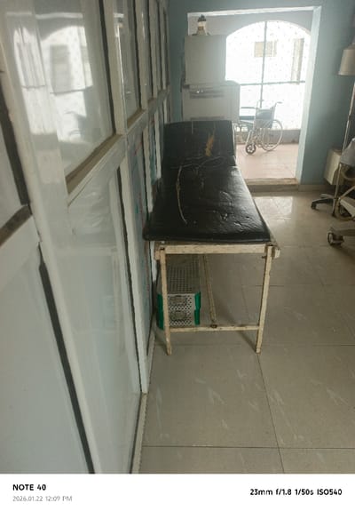The scene depicts an interior space, likely a medical examination room, located in Gwammaja, Nigeria. A prominent black examination couch with a light-colored metal frame occupies the center-right. Its surface is notably worn, exhibiting cracks, peeling, and a distinct yellow discoloration on the upper left section, with a thin dark cord resting on it. Beneath the couch, a silver, perforated medical tray rests on green supports. To the left, a partition composed of multiple white-framed, reflective panels is visible. An arched doorway in the background opens to an outdoor area where a white manual wheelchair is positioned. A metal security gate is visible within the doorway. Immediately inside the doorway, on the right, two white medical devices are stacked, the lower unit featuring a vented design, and the upper unit topped with a yellow-capped bottle. The room features light blue walls and light-colored tiled flooring. A portion of a lamp stand and another medical unit are partially visible on the far right. No individuals are present in the image.
