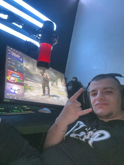 The image displays an indoor gaming setup. The exact location within Blankenberge, Belgium, is not ascertainable from visual information alone.
An adult male subject is prominently featured on the right side of the frame, captured from the chest up and looking towards the camera. He has short dark hair, a visible goatee, and is wearing black over-ear headphones and a dark t-shirt with a white graphic design on the front. His left hand is raised in a "rock on" gesture, with a silver-colored chain bracelet visible on his wrist.
To the left of the subject, a large display monitor presents a video game interface, showing a character in an urban environment and various user interface elements, including "ROUND WINS" and "BUDAPEST." A mechanical keyboard with green and yellow backlighting and a white gaming mouse are situated on a dark surface in front of the monitor. Above the monitor, a red-illuminated condenser microphone is visible, suspended from an adjustable arm. A dark statue, resembling a "Halo" character, is partially visible in the background to the left of the male subject's head. The room is artificially lit, primarily by three parallel horizontal light bars emitting a cool white/blue light from the upper left, complementing the illuminated gaming equipment.