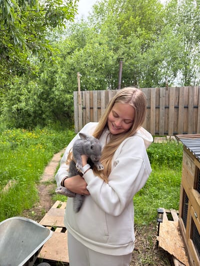 A young woman with long, light brown hair, wearing a white hooded sweatshirt and matching white pants, is captured outdoors, smiling broadly with her eyes closed. She holds a small, fluffy grey rabbit in her arms, looking down at it with evident affection. A white smartwatch is visible on her left wrist, and a black bracelet on her right. The rabbit appears calm and nestled against her.

The scene unfolds in a lush green environment, consistent with a rural or semi-rural setting in Vozhoiskii, Russia. The background is filled with various green trees and bushes, suggesting a daytime shot under an overcast sky. To the right, a weathered wooden fence or a series of wooden structures, possibly animal enclosures, stands against the natural backdrop. A metal gutter pipe runs horizontally from one of these structures. The ground is carpeted with green grass interspersed with small yellow flowers, and a narrow dirt or gravel path is visible winding through the greenery. Partially visible in the lower right corner is a silver-colored metal object, resembling part of a wheelbarrow, next to a small black solar-powered light device on the ground. No text is visible in the image.