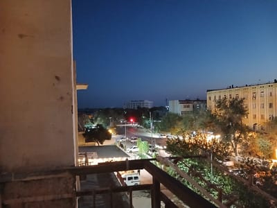 This image captures a street scene at dusk in Qorowul, Uzbekistan. In the foreground, a textured, light-colored wall occupies the left side of the frame, with a metal railing extending horizontally across the bottom. Beyond the railing, a street is visible, illuminated by streetlights and the headlights of vehicles. A traffic light shows a red signal. Several cars and vans are present on the road, some appearing to be parked.
In the middle ground, trees with lush green foliage line the street, their branches illuminated by artificial lights. A few buildings are visible in the background, including a multi-story structure with numerous windows and what appears to be a large, modern building further in the distance. The sky is a deep, clear blue, indicating either early evening or late twilight. The overall impression is one of urban activity at the transition from day to night. There are no discernible individuals clearly visible and identifiable in the scene.