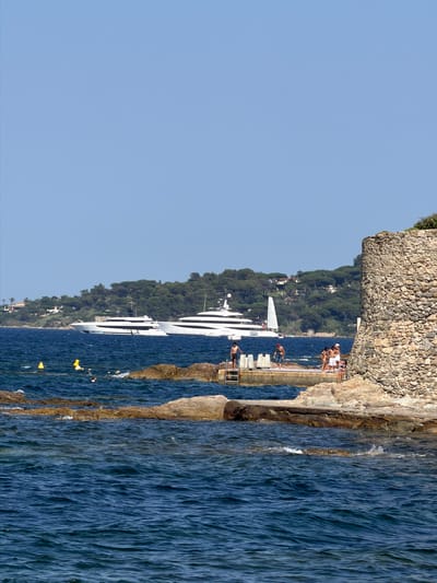 This media file captures a vibrant scene at Saint-Tropez, France, likely during a warm, sunny day given the clear blue sky and people in swimwear. The image is oriented vertically, with the bottom of the frame depicting a vast expanse of deep blue ocean, its surface rippling and catching the light. Along the right edge of the frame, a rocky coastline juts out, with rough, textured stones forming a natural barrier.

On a man-made stone walkway or pier extending from the coastline, several individuals are visible. Some are casually standing and observing the surroundings, while others are engaged in activities near the water. One person is perched on the edge of the walkway, appearing to be preparing to dive or jump into the sea. Another individual is seen in the water, further out. A large, modern white yacht with numerous decks and windows is anchored in the bay, its sleek design contrasting with the natural landscape.

Behind the yacht and the water, a lush green hillside is covered in dense trees, dotted with glimpses of buildings, suggesting a settlement or resort nestled into the landscape. A stone wall, built with rough-hewn blocks, forms the upper edge of the scene on the left. Yellow buoys are scattered across the water's surface, marking an area or possibly part of the yacht's mooring. The overall atmosphere appears relaxed and leisurely, indicative of a vacation setting where people are enjoying the sun, sea, and luxury of a superyacht. No visible text is present in the image.