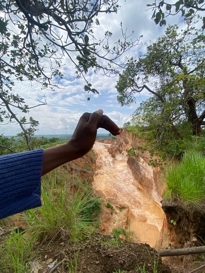 In Amangwu, Nigeria, a hand wearing a blue knitted garment extends into the frame, its index finger and thumb forming a circular gesture over a dry, eroded landscape. The terrain features steep, sandy cliffs with visible layers of sediment, suggesting a former riverbed or quarry. Lush green grass and foliage grow along the edges of the cliffs and in the foreground. In the background, a vast expanse of greenery stretches to the horizon under a sky filled with scattered white clouds. A large tree with dense foliage is visible on the right side of the frame. The overall scene depicts a natural environment undergoing erosion, with a human element interacting with the landscape.