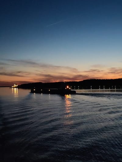 The image depicts a twilight scene over a body of water. The sky transitions from deep blue overhead to vibrant hues of orange, red, and pink at the horizon, indicative of a sunset. A faint aircraft contrail is visible in the upper right quadrant.

A dark, low-profile vessel, likely a barge, traverses the midground water from left to right, its stern lights casting bright yellow reflections on the water and generating a visible wake. On the distant left shore, a prominent, brightly illuminated structure, possibly a building or navigational aid, stands on a small land extension, its yellow lights reflecting on the water's surface. The far shoreline, appearing as a dark, tree-lined landmass, features a series of regularly spaced, vertical white lights extending across the horizon. The foreground water exhibits subtle ripples. No individuals are visible. This scene is located in the city of Yasenevo, Russia.