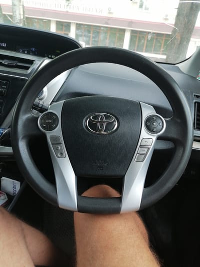 A close-up, eye-level shot shows the interior of a car, specifically the steering wheel and dashboard, from the driver's perspective. The steering wheel, centrally located, features the Toyota logo and various control buttons. The dashboard displays a digital clock reading "1153" and other indicators. Outside the car, through the windshield, a building with multiple windows and a tree are visible. The image appears to be taken in the city of Lesozavodsk, Russia, based on contextual information not present in the image itself.