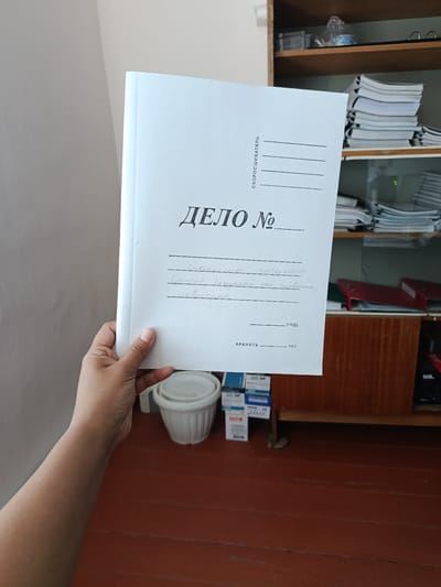 A person's left hand holds a white folder with Russian text. The text on the folder reads "ДЕЛО №" (File No.) and "СКОРОСШИВАТЕЛЬ" (Binder). Below the file number, there is handwritten text which appears to be a description of the contents or purpose of the file. There are also lines for a year and the word "ХРАНИТЬ" (Store).
The background shows a room with wooden flooring and a wooden cabinet with shelves. The shelves are stocked with files, binders, and other office supplies. A white plastic bucket or pot is visible on the floor. The scene is illuminated by natural light, casting shadows. The image appears to have been taken indoors, possibly in an office or storage area. The location is specified as Baetov, Kyrgyzstan.