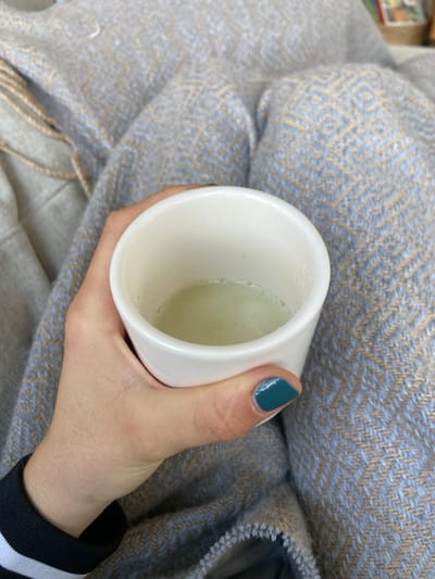 A person's hand, wearing a black and white striped sleeve, is holding a white mug filled with a light green liquid. The hand has teal nail polish on the ring finger. The mug is positioned against a light brown and blue patterned blanket. The coordinates provided (40.952, -73.708) correspond to a location in Rye, New York, United States.