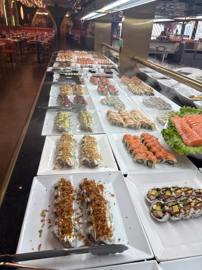 An indoor buffet-style sushi restaurant in Amora, Portugal, displays an extensive selection of sushi and sashimi. In the foreground, several white platters showcase various sushi rolls: one contains rolls topped with crispy fried onions and mayonnaise, another features rolls with avocado and a drizzle of sauce. Adjacent to these, a platter holds salmon sashimi arranged on a bed of lettuce. Further back, multiple rows of sushi rolls, some with colorful toppings and drizzles, are presented on tiered white platters. The buffet features a bright, overhead lighting system that illuminates the food. In the background, the restaurant's dining area is visible with red upholstered seating and intricate light fixtures. There are no people visible in the immediate vicinity of the buffet. The overall environment is clean and well-organized, suggesting a high-traffic dining establishment.