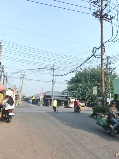 The image shows a street scene in Barengkrajan, Indonesia. The road is asphalt and appears to be a two-lane road with a slight incline. On either side of the road are various businesses and buildings, including what appear to be small shops and stalls. There are several utility poles with a complex network of overhead wires crisscrossing the sky. The sky is clear and blue, suggesting a daytime scene.

Several people are visible in the image, primarily on motorcycles. In the foreground on the left, a person is riding on the back of a motorcycle, wearing a black hijab. On the right side of the road, a person on a motorcycle is wearing a gray long-sleeved shirt and jeans, with a large green woven basket attached to the back of the motorcycle. Further down the road, several other motorcycles are parked or in motion. There are also a few pedestrians, including a person in a bright yellow vest holding a red traffic cone, and another person in a similar yellow vest walking further down the street. The businesses appear to be open, with some displaying signage and awnings. A large, leafy green tree is prominent in the background on the right side of the street. The overall impression is of a busy, commercial street in a relatively modest urban or semi-urban area.