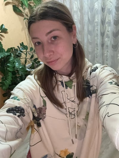 A young woman is captured in a close-up, selfie-style portrait, looking directly at the camera with a slight, pleasant smile. Her head is tilted, and her long, brown hair frames her face, with some strands falling over her shoulder. She has light-colored eyes and fair skin.
She is wearing a cream or off-white hoodie adorned with a detailed floral and vine pattern, featuring black lines and accents of blue, purple, and yellow or orange flowers. White drawstrings with silver metallic tips are visible at the neckline of her hoodie. A simple silver hoop earring can be seen on her right ear.
The setting appears to be indoors in Krapivka, Russia. Lush green potted plants are visible in the foreground and along the lower edge of the frame, adding a natural element to the scene. The background behind her is a textured, light-colored surface, possibly a wall or curtain with a subtle patterned design. The surface below her head is a plain, light yellow or beige. The lighting suggests it is daytime. There is no visible text in the image.