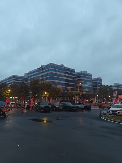 The image captures a street scene in Nanhai District, China, during what appears to be dusk or overcast weather. In the background, a large, modern building complex with blue-tinted windows dominates the skyline. Signs on the building indicate "Global Hardware & Building Materials Manufacturing Base" in English and Chinese. The street in the foreground is wet, suggesting recent rain. Several cars are parked or driving on the road, including a white Buick with license plate "KY 70893" on the right side of the frame. Pedestrians are visible, some with umbrellas, and a person on a scooter is on the left. Red banners are displayed along the street. The overall atmosphere is urban and functional, with a focus on commercial activity.