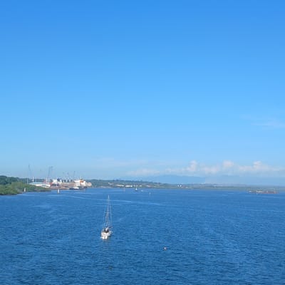 The image presents a wide view of Tanga Bay, Tanzania, under a clear blue sky. The upper portion of the frame is occupied by the bright, cloudless sky. The lower section features blue, rippled water.
A white, single-masted sailboat with two visible outboard motors is situated in the mid-right of the bay, appearing stationary. Several white and orange navigational buoys are scattered across the water. To the immediate left of the sailboat, two small, dark, indistinct objects are visible on the water's surface.
Along the distant left shore, the Tanga Port is operational, featuring multiple large cargo ships docked alongside piers. Numerous tall, grey gantry cranes are positioned over the vessels, suggesting active loading or unloading. Industrial buildings and green coastal vegetation, likely mangroves, line this shoreline.
The distant horizon extends across the frame, showing a continuous landmass with scattered urban development. Faint mountain ranges are visible in the far background, partially obscured by atmospheric haze and topped by sparse white clouds. A large, dark and red cargo vessel or barge is present on the water in the distant right, with other small, indistinct vessels dotting the mid-distance.