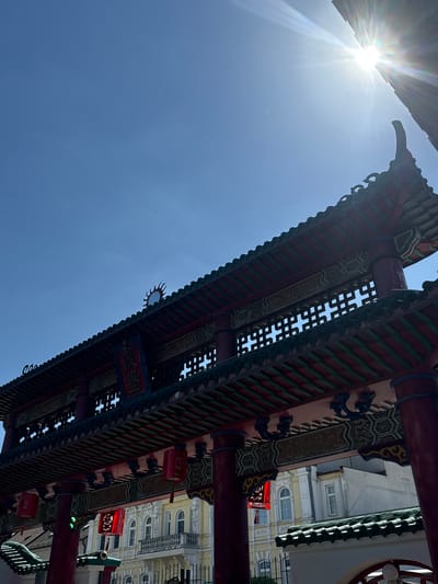 This upward-angled photograph captures a large, ornate Chinese-style gate (Paifang) against a bright blue sky. The gate features traditional architecture with prominent red pillars, intricate green and red tiled roof structures, and decorative elements. Numerous red lanterns with delicate patterns hang from the gate's beams.
The sun is visible in the upper left corner, casting a strong flare and radiating beams of light across the clear sky, indicating bright daylight, likely midday or early afternoon. Tiny dust particles are illuminated by the strong sunlight in the sky.
Through the intricate lattice work and gaps in the gate's structure, parts of a light-colored, multi-story European-style building are visible in the background, featuring arched windows and decorative facades.
Visible text on a red plaque on the gate includes two Chinese characters, which appear to be "廣榮" (Guǎng Róng), often meaning "extensive glory" or similar auspicious connotations.
The scene depicts a unique cultural landmark under a clear, sunny sky, creating a contrast between the ornate Chinese gate and the underlying European urban architecture. Given the location hint, this image is from Hainburg an der Donau, Austria.