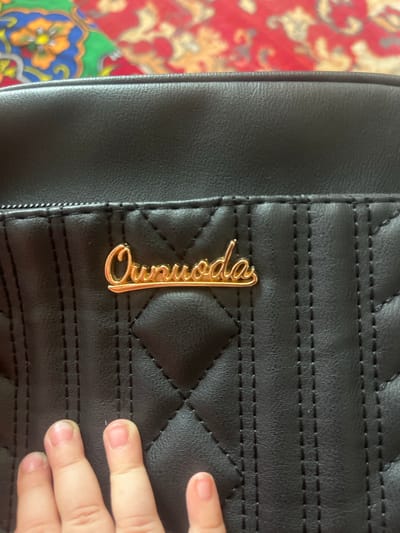 A close-up shot focuses on the quilted black exterior of a handbag. A gold-colored metal logo, reading "Ournoda," is affixed to the bag's surface. The handbag is placed on a vibrantly colored textile with a red and blue floral pattern. The bottom left corner of the image shows the tips of several fingers, suggesting a person is holding or interacting with the bag. The scene appears to be indoors, possibly in a home, given the presence of the handbag and the patterned textile which resembles a rug or tablecloth. The specific location of Ташлак, Kyrgyzstan is not visually evident from the image.