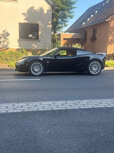 A black convertible Lotus Elise is parked on the right side of a two-lane asphalt road, with a white line marking the center. The car is angled slightly towards the viewer. A person wearing a yellow shirt and glasses is visible in the driver's seat. To the left of the road, there is a light-colored building with a window displaying a "Verkauft" (Sold) sign. Further back on the left, there is a green, leafy hedge. To the right of the road, a red brick building with a tiled roof and dormer windows is visible. The sky is clear and blue. The scene appears to be in a residential area on a sunny day. The location is identified as Bonn, Germany.