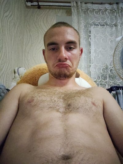 A close-up, head-on, selfie-style shot shows a man with short hair and a light beard looking directly at the camera with a pouty expression. He is shirtless, revealing a hairy chest and torso. Behind him, a stuffed animal is visible, along with a patterned wallpaper and lacy curtains. A fan can be seen on the right side of the frame. The image appears to have been taken indoors. No specific location in Okhtyrka, Ukraine can be determined from the provided image.