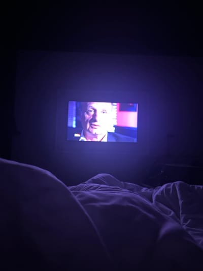 In a dark room, a TV screen displays a man's face, tilted slightly. The screen is mounted on a wall, with a frame around it. To the right, bedding material is visible, possibly a duvet or pillow. The lighting casts a predominantly blue and purple hue over the entire scene. The darkness and orientation suggest the viewer may be in bed.