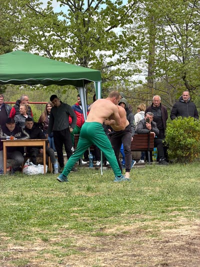 The image captures a scene outdoors, likely in a park in Dulevo, Bulgaria, given the context. Two shirtless men are wrestling, their bodies intertwined in a grappling posture. They stand on a patch of grass, which transitions into a drier, more barren area in the background. A crowd of onlookers surrounds the wrestlers, some seated on benches and chairs, others standing. A green awning or tent structure is partially visible on the left side of the image, offering some shade to the spectators. Various objects are strewn around the scene, including water bottles and what appears to be a trash bag near a wooden table. The environment features trees with green leaves and a generally natural setting. The interactions between the men wrestling and the observers create a sense of an event or competition taking place in a public space.