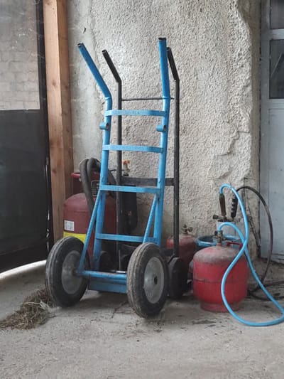 The image shows a blue metal cart with two wheels, possibly a dolly or hand truck, parked next to a red air compressor tank. The cart has black handles at the top. The air compressor tank is round and cylindrical, and a blue hose is connected to it. It appears to be an outdoor or workshop setting, with a rough textured wall in the background and a wooden shelf or beam below the cart. The lighting suggests it is daytime. There is some dry grass or straw on the ground near the wheels. The scene is static, with no activity taking place. There is no visible text in the image. The location is identified as Kochmar, Bulgaria.