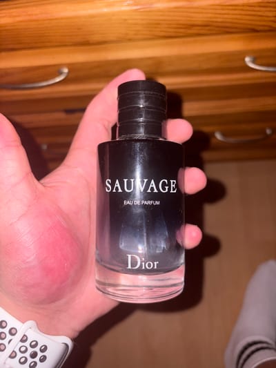 A hand is holding a nearly empty bottle of Dior Sauvage Eau de Parfum. The hand has a white and black smartwatch on the wrist. The background includes a wooden cabinet with silver handles and a person's feet with white socks and black shoes. The location is not specified in the image.