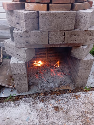 An outdoor scene depicts a makeshift fireplace constructed from cinder blocks. The structure is roughly built, with blocks stacked unevenly to form an opening in the front. Inside this opening, a fire burns, casting a warm orange glow. Embers and ash are visible at the bottom of the fire chamber. The surrounding ground appears to be a mix of concrete and dirt, with scattered leaves and debris. The background shows a portion of a light-colored building, possibly a house or shed, with vertical siding. The overall impression is of a rustic, functional outdoor fireplace. The location is identified as Vysokaya Gora, Russia.