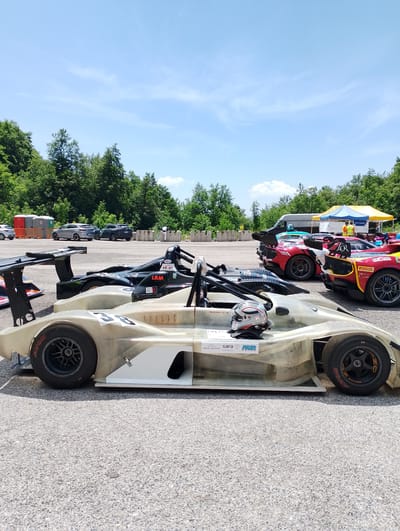 The image depicts a row of open-wheel race cars parked on an asphalt surface, likely a racetrack or pit lane, under a bright, partly cloudy sky. In the foreground, a light-colored, unfinished race car with the number "38" is prominent. A racing helmet rests on its cockpit. Behind it, several other race cars are visible, including a black car with "LAM" and "BELVA" logos, and a red and yellow car with "REOFER" and "PIRELLI" branding. In the background, a yellow and blue tent is set up, with several people milling about. Beyond the track, a dense line of green trees borders the area. The presence of portable toilets and barrier walls suggests this is a temporary setup, common for racing events. The overall atmosphere suggests a racing event is either about to begin or has just concluded. The context provided about Rieti, Italy, places this scene in an outdoor, possibly rural or semi-urban setting within the country, characteristic of motorsport venues.