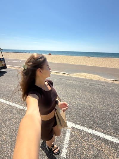 A woman is walking on a paved path beside a beach. The sky is clear blue, and the sea is visible in the background. The woman is wearing a brown crop top and matching shorts, and she has a shopping bag in her hand. She is also wearing black sandals. There is a vehicle on the left side of the image, and some beach huts are visible in the distance on the beach. The scene appears to be a sunny day in Bournemouth, United Kingdom.