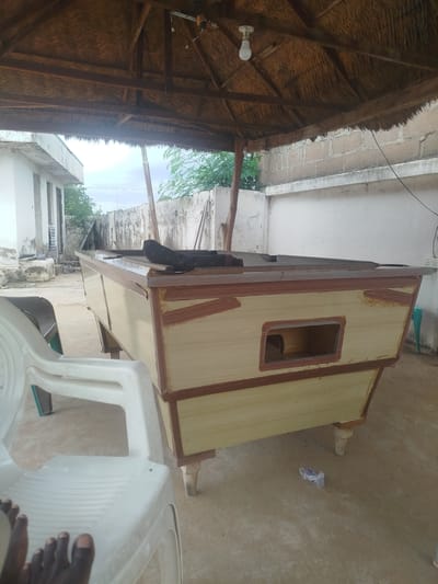 An outdoor scene under a thatched roof structure features a partially constructed wooden pool table. The pool table is light yellow with brown trim and has a rectangular opening on its side. A black cloth is draped over a portion of the pool table's surface. In the foreground, a white plastic chair is visible with a person's feet resting on it. The ground is sandy and appears to be a dirt floor. In the background, there is a light-colored building with a rough texture and some vegetation. The sky is overcast. The scene suggests a casual outdoor setting, possibly a workshop or recreational area in Yola, Nigeria.