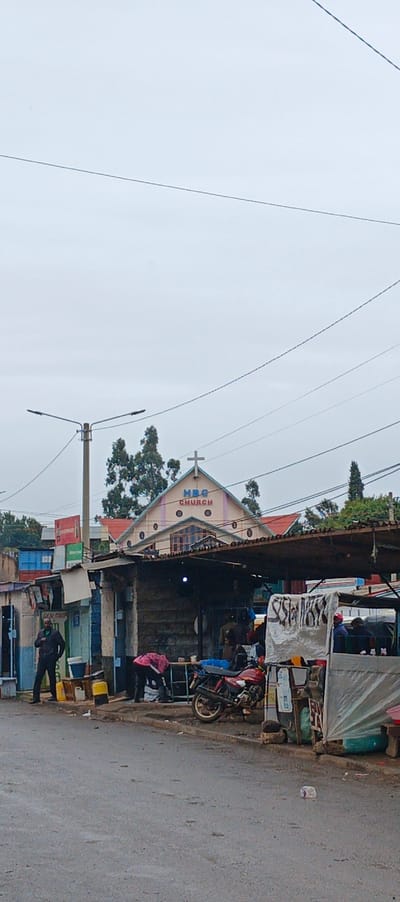 A street scene features a small church in the background with "HBG CHURCH" displayed above a cross on its roof. Below, the foreground is filled with informal roadside businesses under corrugated iron roofs. People are present, some standing near the storefronts and another bending over at their work. A red motorcycle is parked among the structures. Power lines crisscross the cloudy sky above. The wet pavement suggests it has been raining recently.