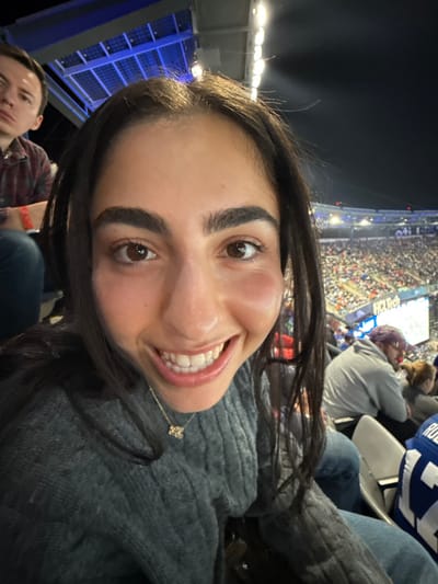 A wide-angle, eye-level photograph captures a young adult female in the immediate foreground, smiling broadly directly into the camera from a stadium seat at night. She has dark hair and wears a grey cable-knit sweater and a gold necklace with a small pendant. Her cheeks exhibit slight distortion from the wide-angle lens.

To her upper left, a young adult male with light skin, wearing a plaid shirt and a red wristband, is partially visible looking upward. To her right and slightly further back, another male spectator in a light grey hooded sweatshirt and a purple cap is seen, with a red object near his mouth. A portion of a blue and white "Giants" jersey is visible on a person seated in the lower right.

The background reveals a large, illuminated stadium bowl filled with numerous spectators. Bright floodlights are prominent above the seating areas. A large video screen displaying "HCLTech" is visible in the distance. Structural elements resembling blue-tinted solar panels are present above the stadium architecture against the dark night sky. This scene is identified as taking place at MetLife Stadium in East Rutherford, New Jersey, United States.