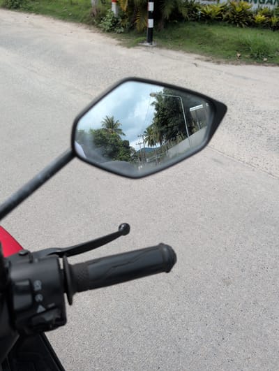The image is taken from the perspective of a rider on a motorcycle traveling on a paved road. The rider's right handgrip and rearview mirror are visible. The rearview mirror reflects a scene of a road lined with trees and utility poles. The reflected image appears to be a road in a populated area with residential or commercial buildings. The road surface is a light gray concrete. The environment suggests a tropical or subtropical climate due to the presence of palm trees. Based on the description, the location is identified as Moo 4, Thailand.