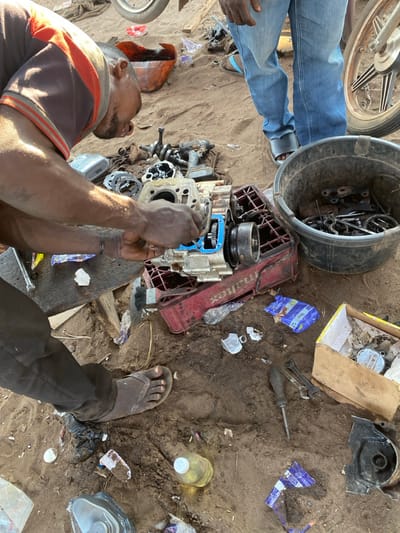 A man repairs a motorcycle engine outdoors, surrounded by tools, parts, and debris on the ground. The engine is partially disassembled, resting on a red crate. A bucket filled with metal parts sits nearby. Another man wearing jeans is standing close to the engine. Discarded containers, plastic wrappers, and tools are scattered on the dirt surface.