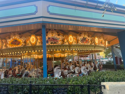 The image captures a brightly lit carousel, likely situated in an amusement park or fairground. The carousel's upper structure features a multi-tiered canopy painted in light blue, cream, and a pale green or yellow, with dark blue trim. Below the canopy, an ornate, gilded frieze is adorned with numerous warm-toned light bulbs and decorative panels.

Several panels are clearly visible:
*   One panel displays a painting of a vintage orange pickup truck.
*   Another panel shows a scene with several American flags and what appears to be a historical tableau, possibly depicting early American settlers or figures.
*   A third visible panel, partially obscured, seems to depict a sailing ship or nautical theme.

The carousel itself is filled with numerous animal figures, predominantly horses, in various poses. Most of the horses are painted in shades of white, brown, or grey, some with elaborate saddles and bridles. The carousel appears to be stationary or has just completed a cycle, as no riders are distinctly visible, and the animals are in their upright positions.

In the immediate foreground, a dense row of green bushes, possibly boxwood or similar foliage, creates a natural barrier. Behind the bushes, a black metal fence with decorative elements is visible. Beyond the carousel, other park structures, trees, and what looks like a large, round satellite dish-like object can be seen in the background.

The lighting suggests it is either late afternoon or early evening, as there is still natural light in the sky and on the surrounding trees, but the carousel's numerous lights are fully illuminated, casting a warm glow. No discernible text is visible anywhere in the image.