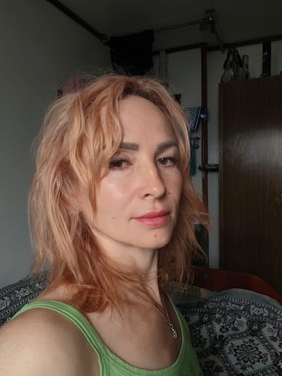A woman with shoulder-length, wavy, light brown hair is shown in a close-up, head-and-shoulders shot. She is wearing a light green tank top and a silver necklace with a small pendant. Her skin is fair with some freckles and a light dusting of makeup on her eyes and lips. She is looking towards the camera with a neutral expression.
The background appears to be an indoor setting, possibly a bedroom or living room. To the right of the woman is a wooden wardrobe or cabinet with a dark wood finish. Above and to the left of the woman, there are elements of the room, including what looks like a ceiling fixture and a grey wall. A patterned blanket or throw is visible on what appears to be a bed or chair behind her.
The image does not provide any specific indicators of the location being in the city of Shalsky, Russia.