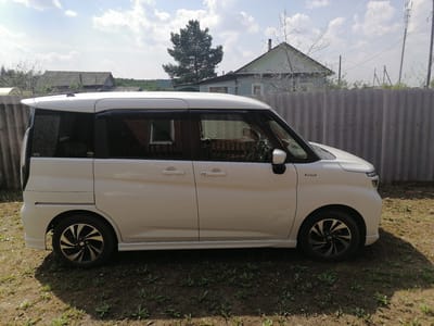 A white, four-door minivan is parked on a patch of dry grass and dirt in front of a grey corrugated metal fence. The minivan's side is facing the camera, and its wheels are visible. The background shows a light blue house with a dark grey roof, a tall evergreen tree, and a cell tower. The sky is partly cloudy and blue. The location is identified as Lobkovo, Russia.