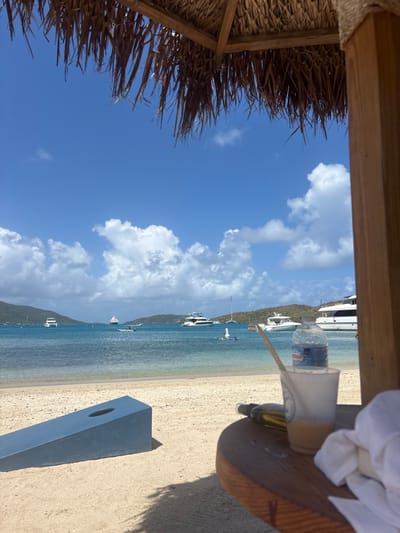 The image depicts a sunny daytime scene on a sandy beach in Spanish Town, British Virgin Islands, viewed from beneath a thatched roof structure with a wooden support post visible on the right.
In the immediate foreground on the right, a circular wooden table displays a clear plastic cup containing a light-colored beverage and a wooden stir stick. A "Crystal Geyser" plastic water bottle rests inside the cup. Adjacent to the cup, a dark-colored bottle lies horizontally, and a crumpled white cloth or towel is present. The thatched roof, composed of dried fronds, overhangs the top portion of the frame, casting a shadow over the foreground.
The mid-ground features a light-colored sandy beach. A blue-painted cornhole board is positioned on the sand, angled towards the water. Beyond the beach, calm, clear turquoise water extends across the frame. Multiple vessels are anchored or moored in the bay, including at least five large white yachts, several smaller boats, and sailboats. A dark-hulled vessel is situated near the center of the bay. Two individuals are present in the water: one appears to be on a white paddleboard or inflatable, while another is partially submerged nearby.
The background consists of green, vegetated hills forming distant shores under a clear blue sky dotted with scattered white cumulus clouds.