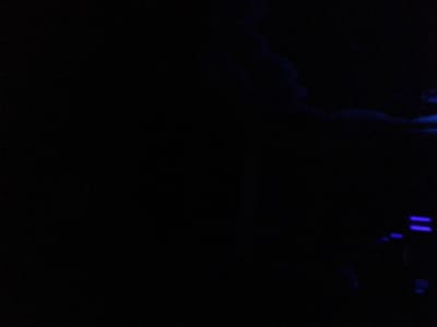 The image displays an extremely dark, low-light indoor environment, consistent with a themed exhibit or attraction, located at the Aquarium of the Pacific in Long Beach, California, United States. The scene is dominated by a pervasive blackness, with illumination provided by faint blue light. In the upper right quadrant, indistinct, wavy blue forms are visible, resembling abstract illuminated elements or reflections within a dark space. The lower right quadrant features at least two distinct, parallel horizontal blue light strips, likely artificial lighting fixtures such as LEDs, along with other diffused blue light sources. No individuals, discernible objects beyond the lighting elements, or specific actions/interactions are identifiable due to the severe lack of light. The environment appears to be a controlled setting designed for mood lighting.