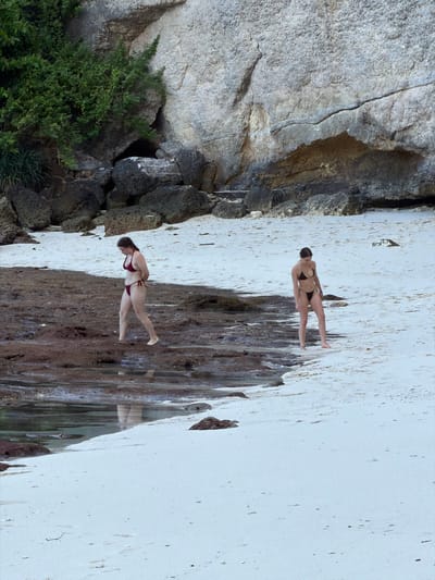 The image shows a coastal scene in Pejukutan, Indonesia. Two women in bikinis are standing on a beach or rocky shore. One woman is wearing a dark-colored bikini and is positioned towards the upper-right of the frame. The other woman, wearing a red bikini, is closer to the bottom-left. The terrain consists of a light-colored sandy or rocky area transitioning into darker, wet rocks, possibly due to wave action or a stream. To the left, there's a rocky cliff or hillside covered with vegetation. The overall composition of the photo is from an elevated perspective, and the orientation is rotated counter-clockwise.