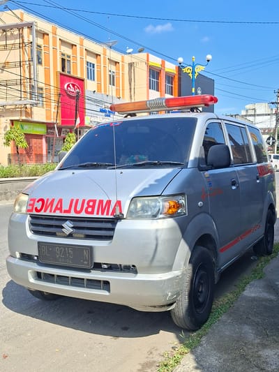 An exterior, eye-level, medium shot captures a silver ambulance parked on the right side of a street in the city of Bireuenmeunasahcapa, Indonesia. The ambulance, a Suzuki APV, is the primary subject. Red lettering on the front of the vehicle reads "AMBULANCE" in reverse. A red and orange light bar sits atop the vehicle's roof. The ambulance features a license plate that reads "BL 9215 N 09-24". A red heartbeat graphic is visible on the side of the vehicle.

The background consists of a two-story building with a beige and orange facade, featuring several windows and a prominent red sign with a white logo. Power lines are strung across the top of the frame. A blue lamppost with yellow accents stands near the building. The street is paved, and a strip of green grass and a concrete curb are visible to the right of the ambulance. The sky is clear and blue with a few white clouds. No people are visible in the frame. The scene appears to be a sunny day.